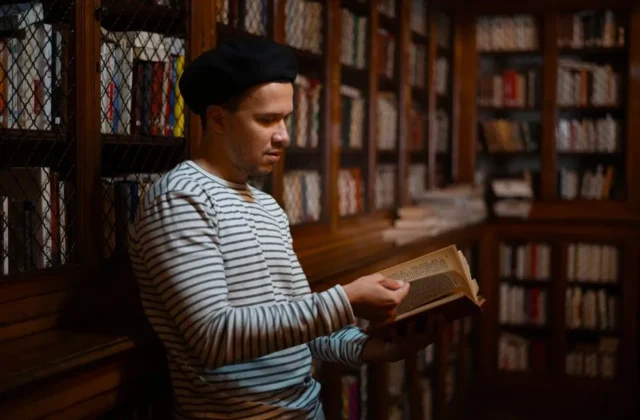 A man in a striped shirt reads a book in a warmly lit library filled with bookshelves.