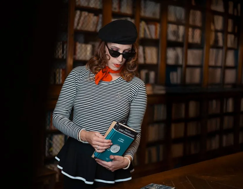 Elegant woman in a striped shirt and beret reading in a classic library setting.