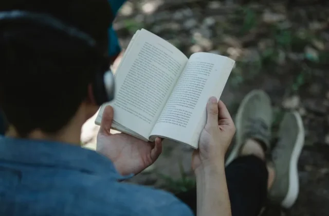 From above back view of crop unrecognizable male listening to music via headphones and reading book while sitting on chair in park