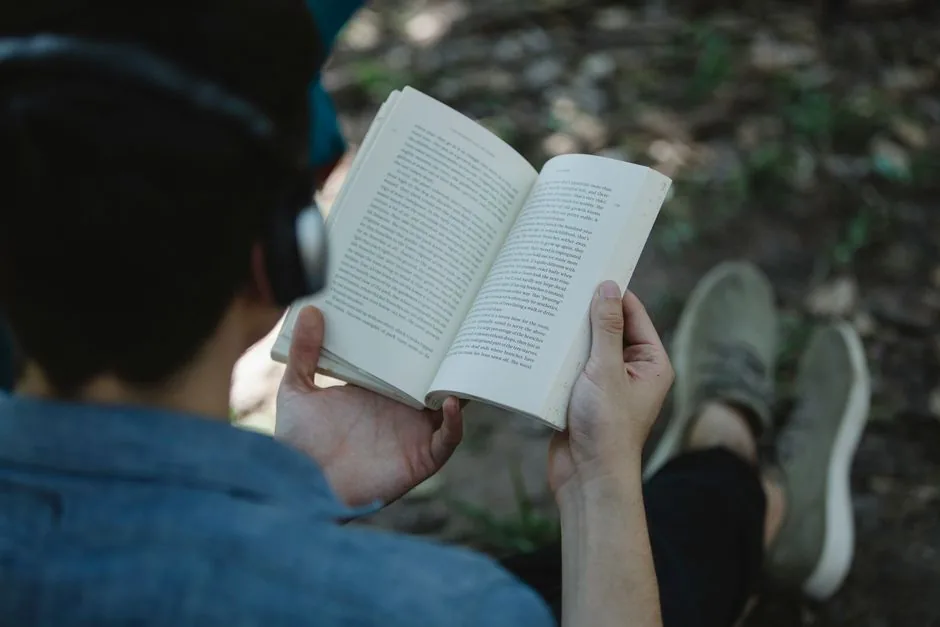 From above back view of crop unrecognizable male listening to music via headphones and reading book while sitting on chair in park