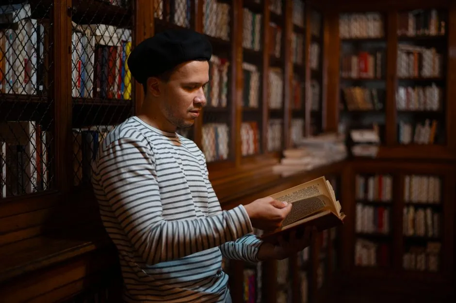 A man in a striped shirt reads a book in a warmly lit library filled with bookshelves.