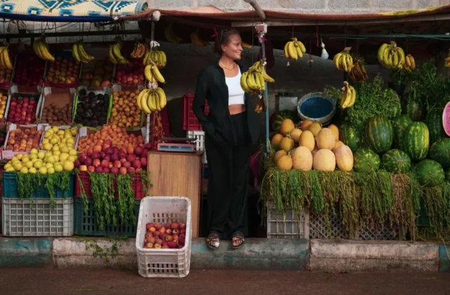 A woman stands by a colorful fruit stall in Oued Ed-Dahab with bananas, melons, and more.