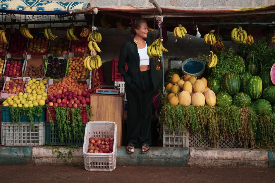 A woman stands by a colorful fruit stall in Oued Ed-Dahab with bananas, melons, and more.